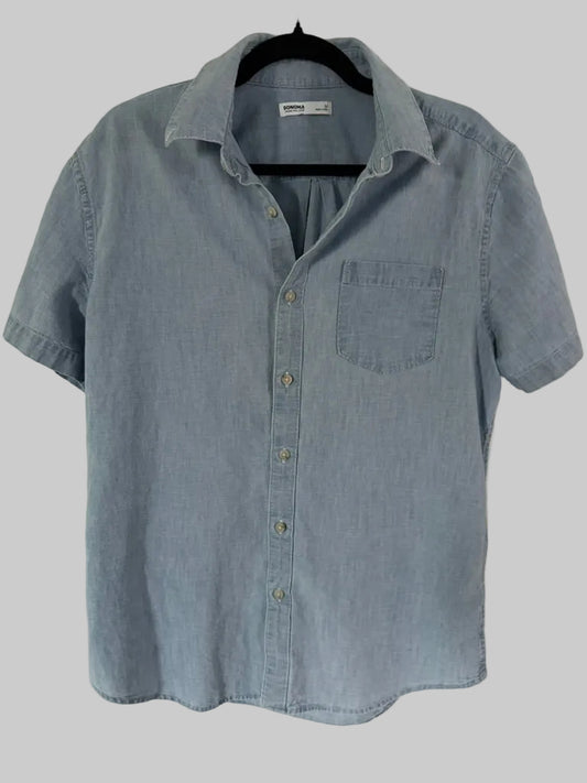 A light blue chambray short-sleeve button-up shirt hanging on a black hanger against a white paneled door.