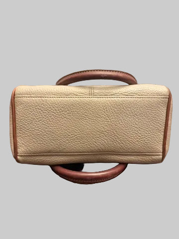 A beige pebbled leather handbag with brown leather trim and handles, resting on a dark wood floor in front of a window with blinds.