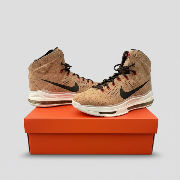 A pair of Nike high-top sneakers with a cork-textured beige upper, white midsoles, and red laces, displayed on top of an orange Nike shoebox against a gray paneled cabinet background.