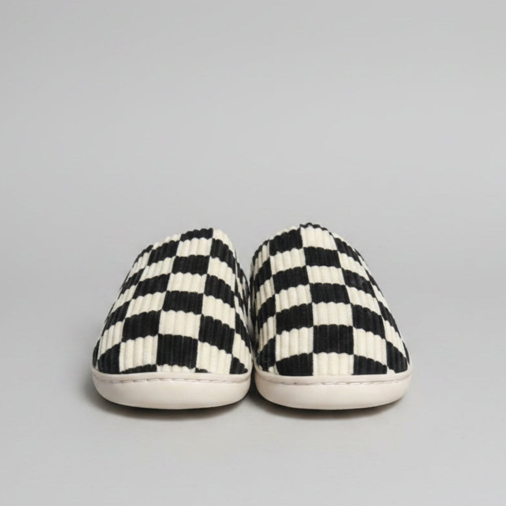 A pair of slip-on slippers with a black and off-white checkerboard pattern and ribbed texture, resting on a gray shag rug.