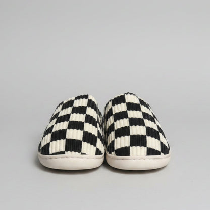A pair of slip-on slippers with a black and off-white checkerboard pattern and ribbed texture, resting on a gray shag rug.
