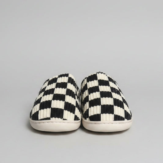 A pair of slip-on slippers with a black and off-white checkerboard pattern and ribbed texture, resting on a gray shag rug.