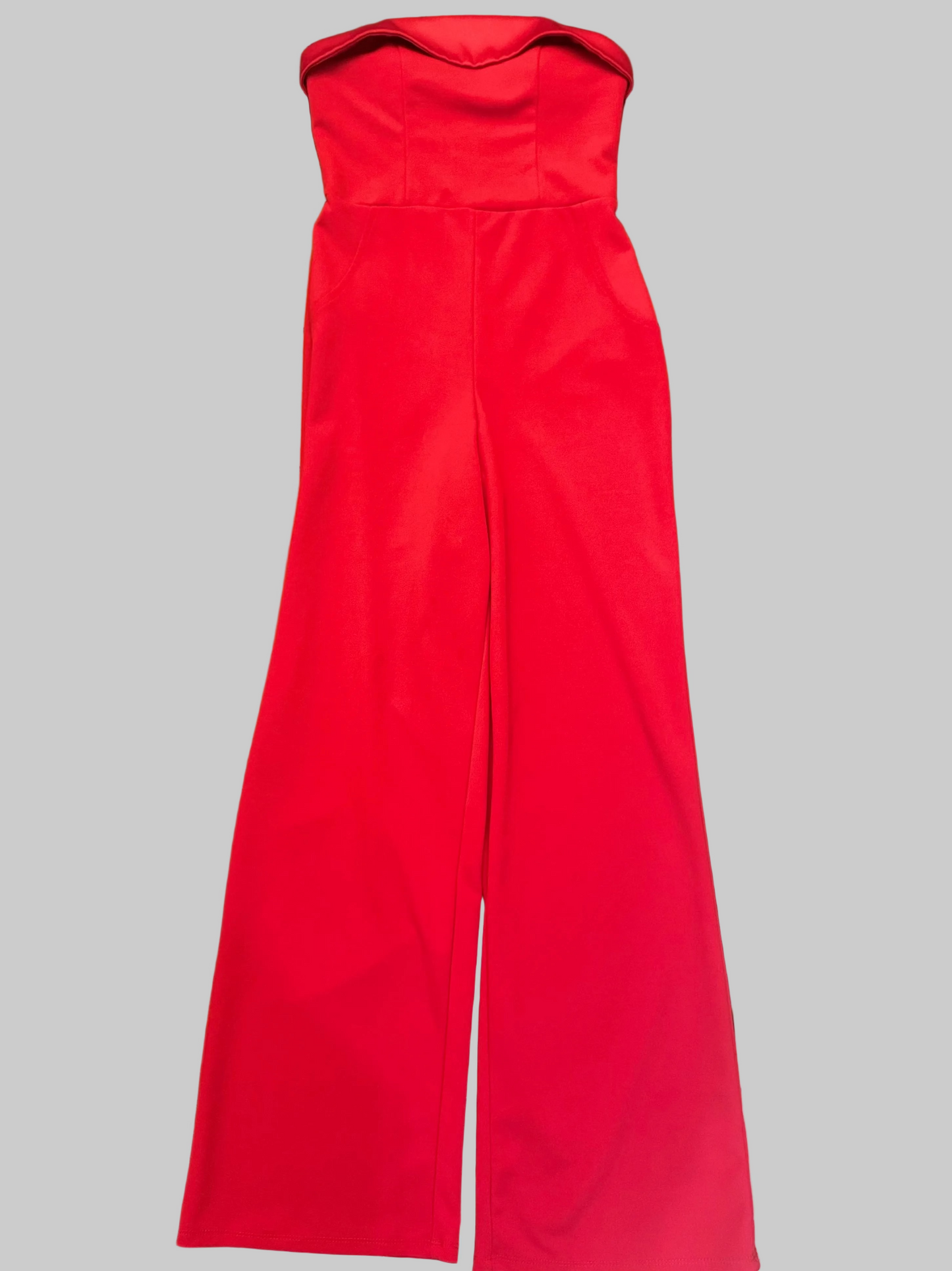 A bright red strapless jumpsuit with wide legs, laid flat on a dark wooden table. The background contains a small round wooden tray with sunglasses and a candle, a blue-handled lint roller, and a tiled floor.