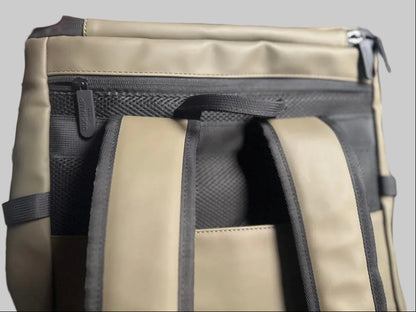 Close-up of a backpack being held open by a hand, showing the interior zippered compartment, mesh lining, and padded straps in a muted olive-tan and black colorway.