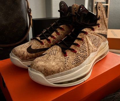 A pair of Nike high-top sneakers with a textured beige and brown cork-like upper, black swooshes, and red laces, resting on an orange shoebox.