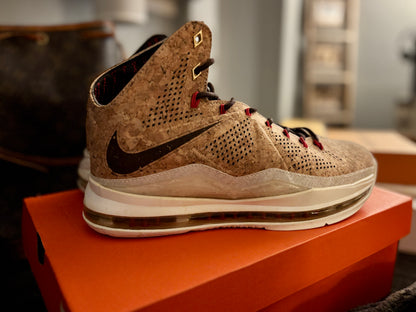 A Nike high-top basketball shoe with a textured tan and brown perforated upper, black Swoosh, and red laces, resting on an orange shoebox.