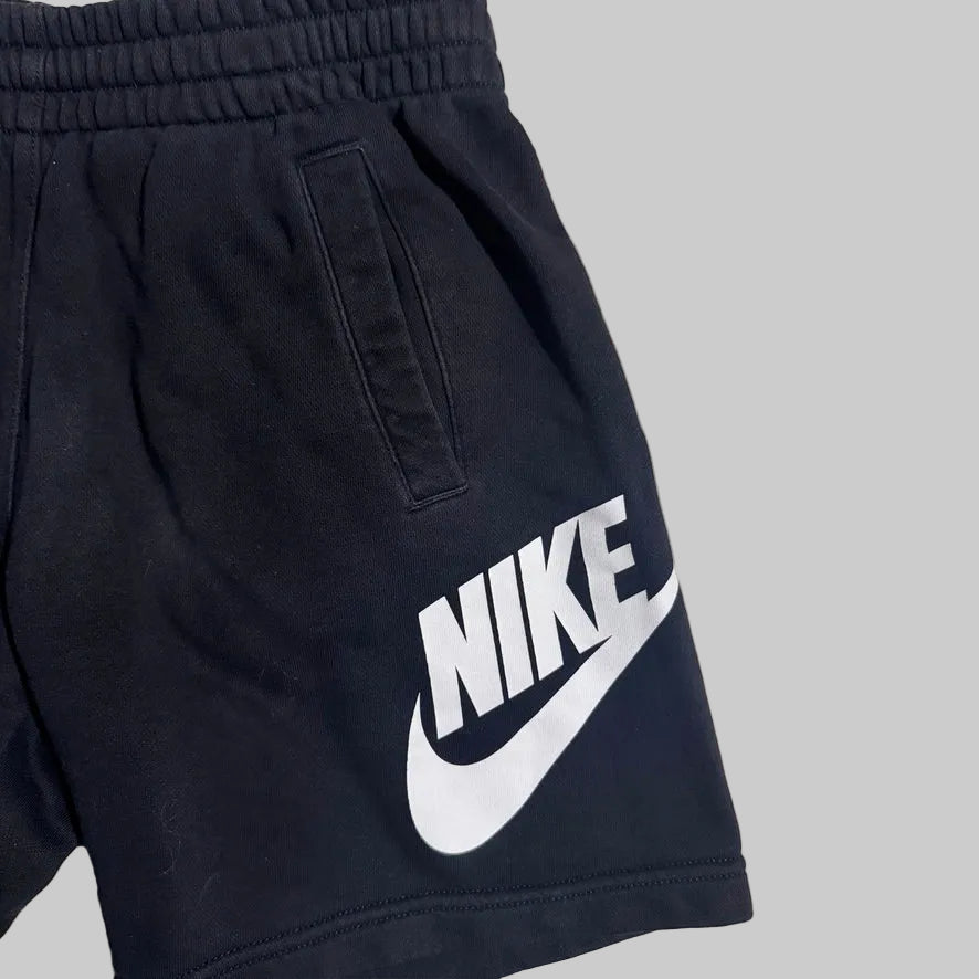 Close-up of black Nike shorts with a large white Nike logo and swoosh printed on the side, shown against a plain light gray background.