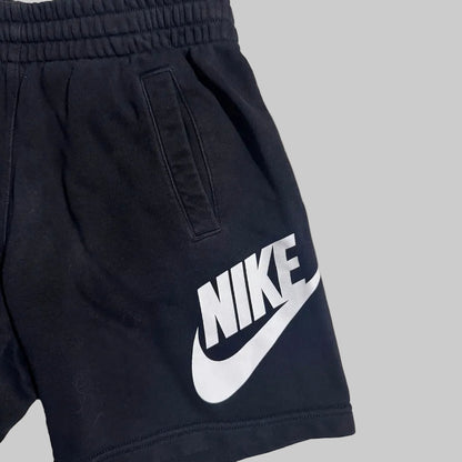 Close-up of black Nike shorts with a large white Nike logo and swoosh printed on the side, shown against a plain light gray background.