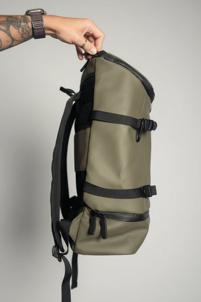 A person's hand is shown holding the top zipper of an olive green waterproof backpack with black straps and zippers, set against a blurred bedroom background.