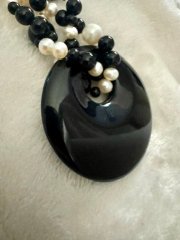 A black beaded necklace with a large, smooth, oval black pendant, resting on a textured white fabric background.
