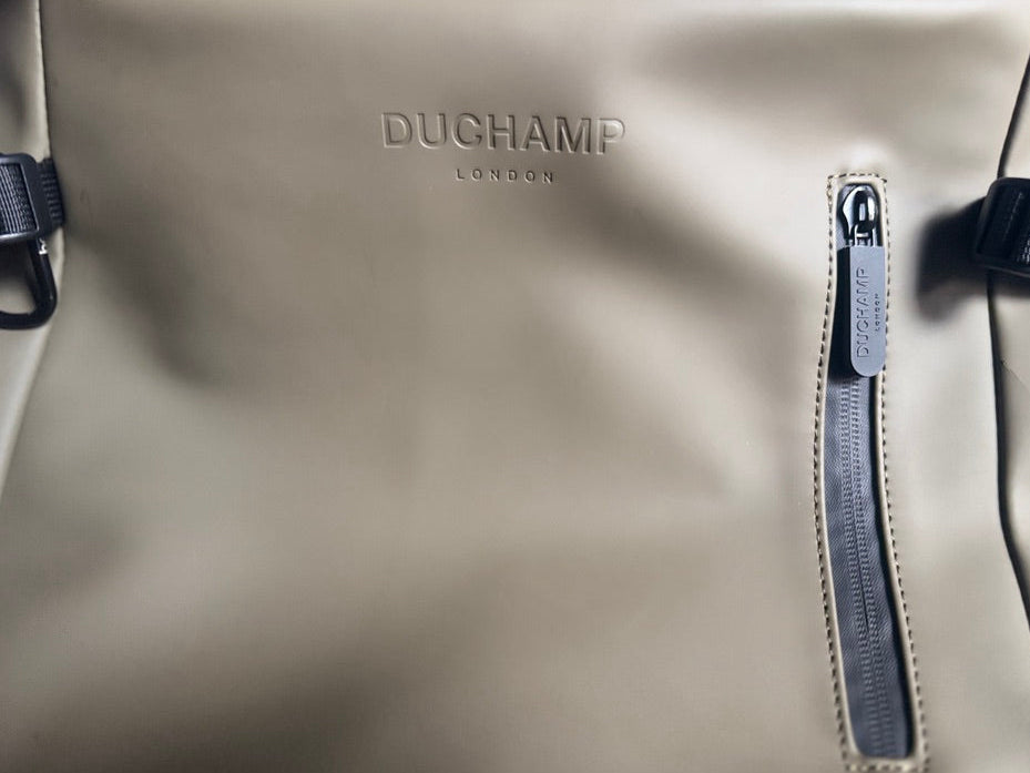 Close-up of a beige DUCHAMP London waterproof backpack with a vertical zipper pocket and black straps.