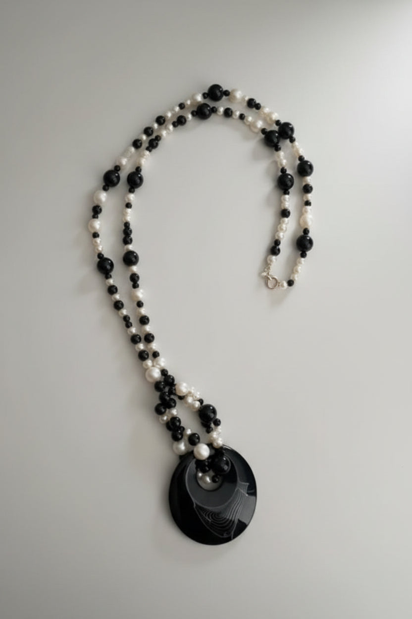 A black and white beaded necklace with a large black circular pendant featuring a crescent moon design, resting on a textured light gray surface.