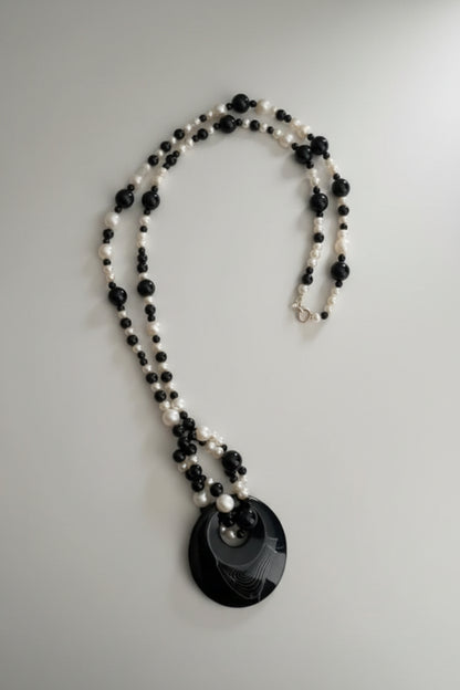 A black and white beaded necklace with a large black circular pendant featuring a crescent moon design, resting on a textured light gray surface.