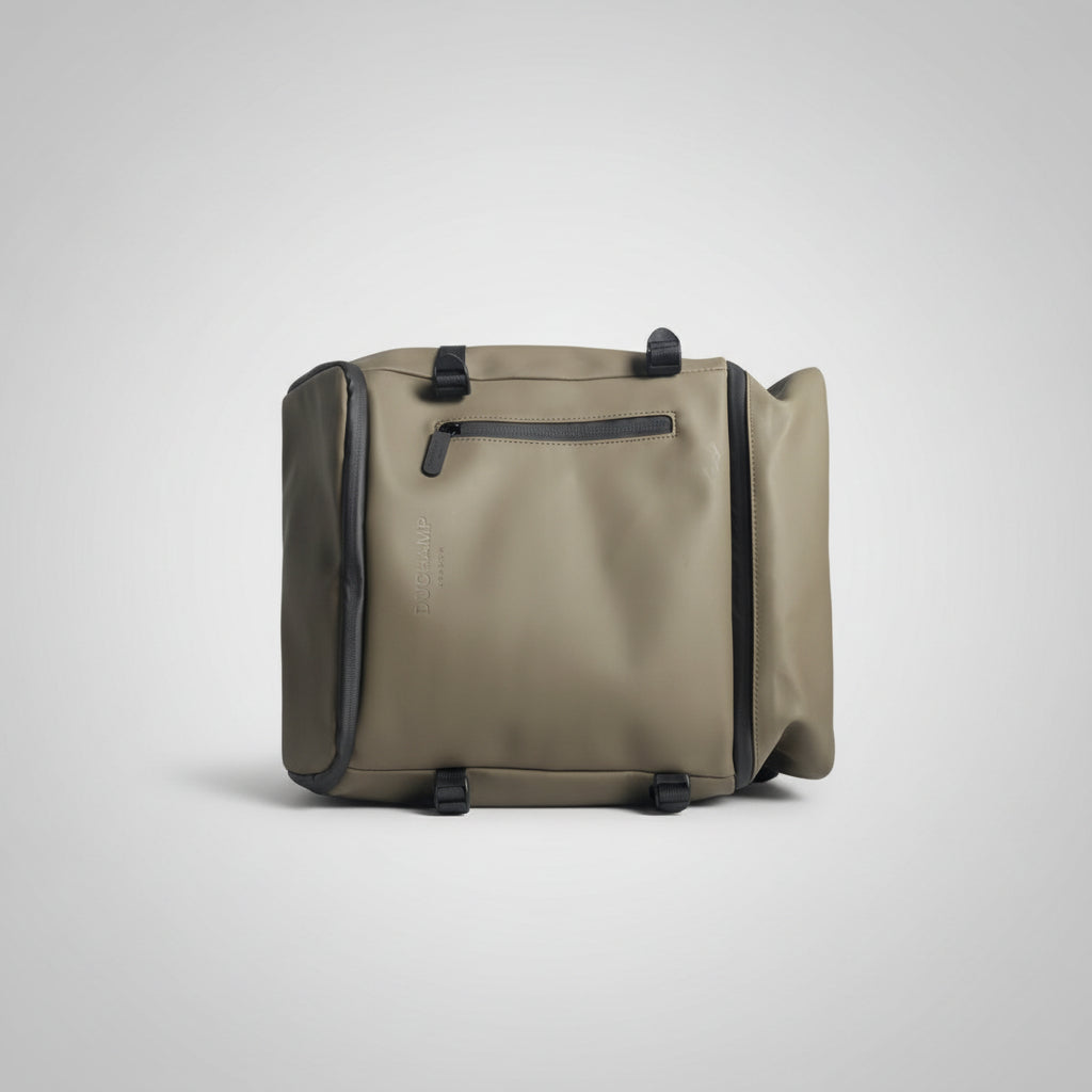 A sage green waterproof duffel bag with black trim and straps, held by a hand in a bedroom setting.