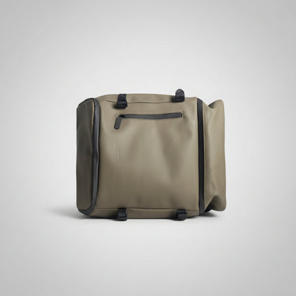 A sage green waterproof duffel bag with black trim and straps, held by a hand in a bedroom setting.