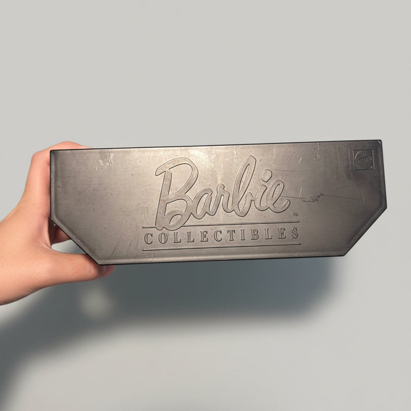 A hand holds a dark gray rectangular metal plaque featuring the embossed Barbie Collectibles logo, set against a blurred background of a blue and white plaid shirt.