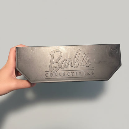 A hand holds a dark gray rectangular metal plaque featuring the embossed Barbie Collectibles logo, set against a blurred background of a blue and white plaid shirt.
