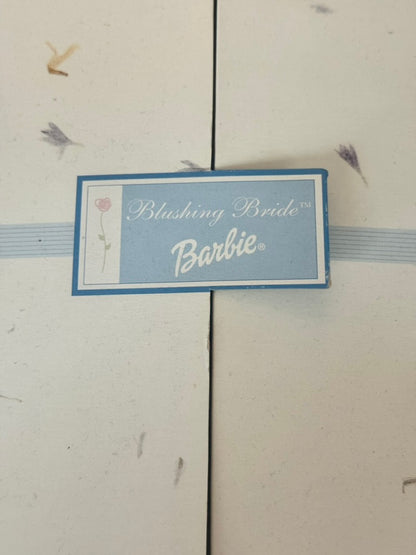 A small rectangular Barbie-themed card or tag reading 'Blushing Bride Barbie' with a light blue background and white script, photographed against a cream-colored surface with a vertical seam.