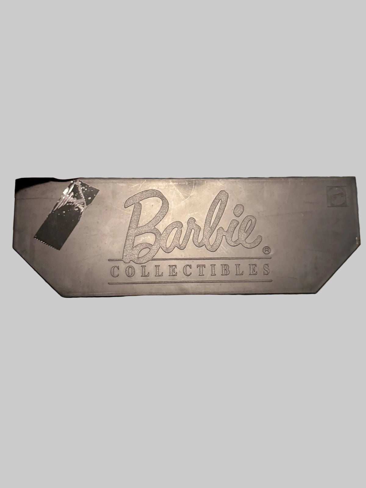 A hand holding a dark gray rectangular collectible plaque featuring the embossed Barbie Collectibles logo, set against a blurred blue and white striped background.