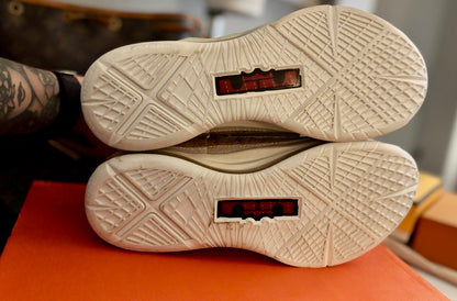 Close-up of the outsoles of a pair of beige athletic shoes, showing textured tread patterns and red translucent heel inserts, resting on an orange surface.