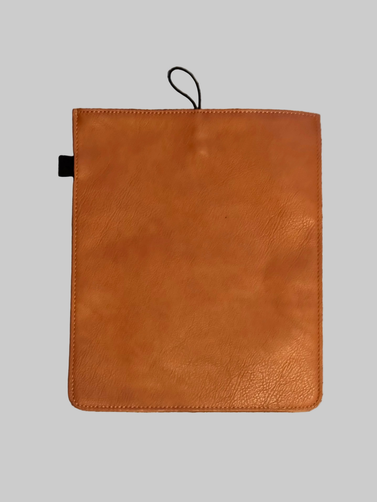 A tan leather tablet sleeve or folio with a black loop tab at the top, laid flat on a gray textured carpet.