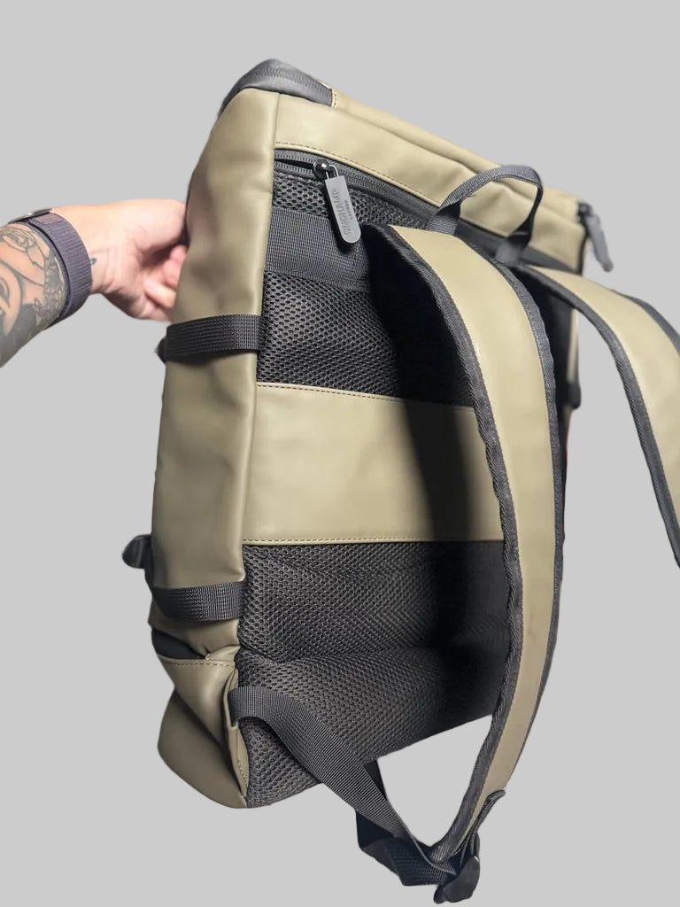 A close-up of a backpack shown from the back, held by a tattooed hand. The backpack features a sage green canvas body with black mesh panels and black straps, set against a blurred bedroom background.