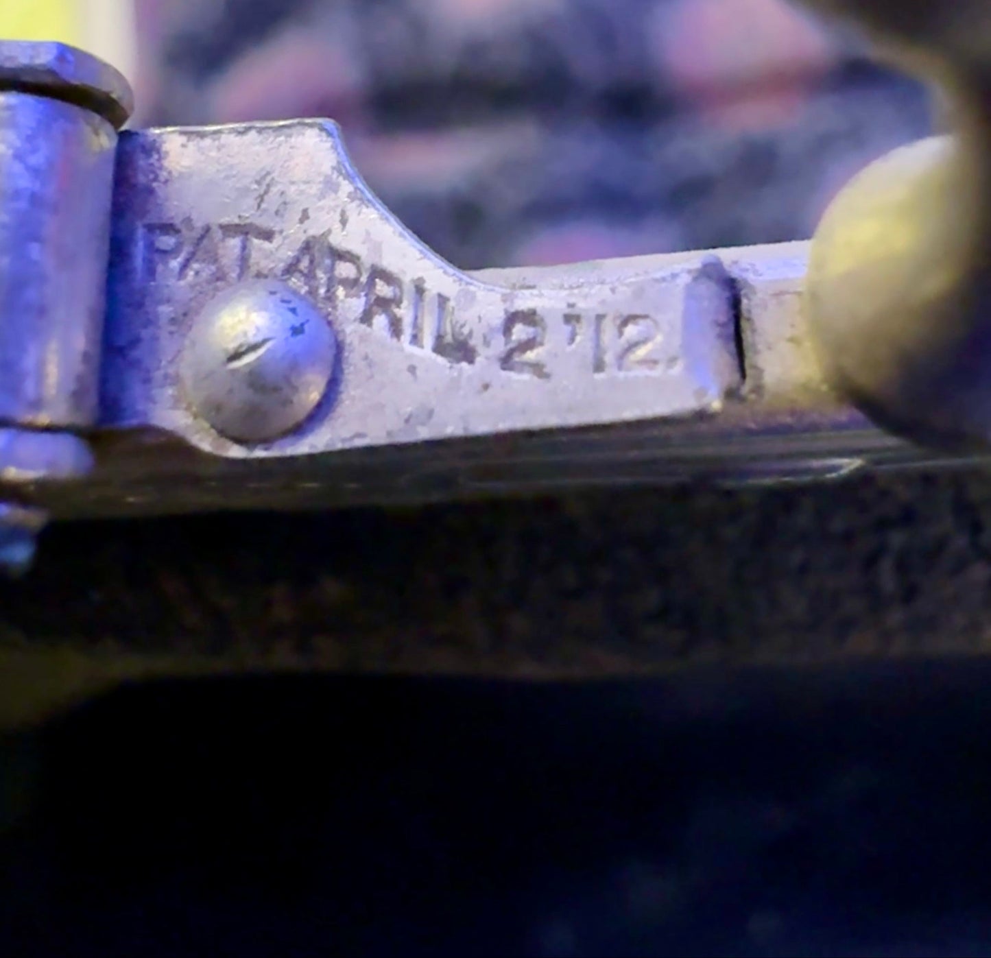 Close-up of a metal object with 'PATENTED' and 'APRIL 2 1912' engraved on it, blurred background