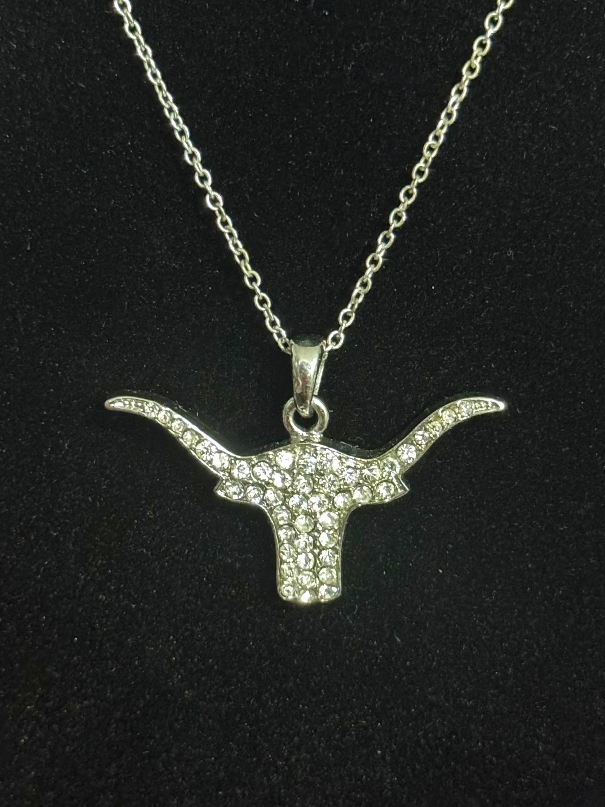 A silver-toned longhorn bull head pendant necklace paved with small clear stones, displayed on a black textured background.