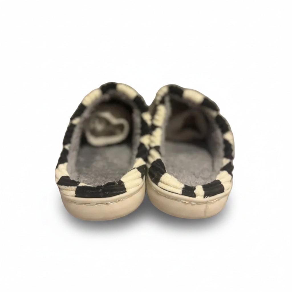 A pair of slip-on slippers with a black and white cow-print patterned upper and a gray plush lining, resting on a gray textured rug.