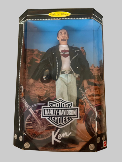 A boxed Ken Harley-Davidson action figure displayed on a light-colored tiled floor. The figure is dressed in a black leather jacket, white t-shirt, and light blue jeans, posed with a motorcycle inside the packaging.