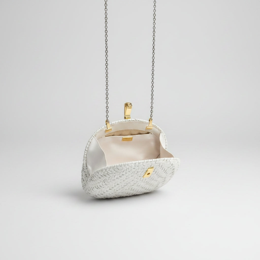 A small structured evening bag with a textured cream exterior, cream lining, and gold-tone hardware, hanging over a patterned rug in an indoor setting.