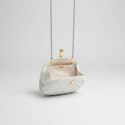 A small structured evening bag with a textured cream exterior, cream lining, and gold-tone hardware, hanging over a patterned rug in an indoor setting.