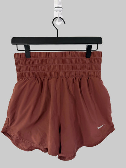 A pair of Nike athletic shorts in a dusty terracotta brown color, hanging from a black hanger against a white paneled door. A small white Nike swoosh logo is visible on the lower left leg.