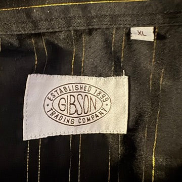 Close-up of a garment's interior label reading GIBSON ESTABLISHED 1999 TRADING COMPANY, with a small size tag marked XL, on a dark pinstriped fabric background.