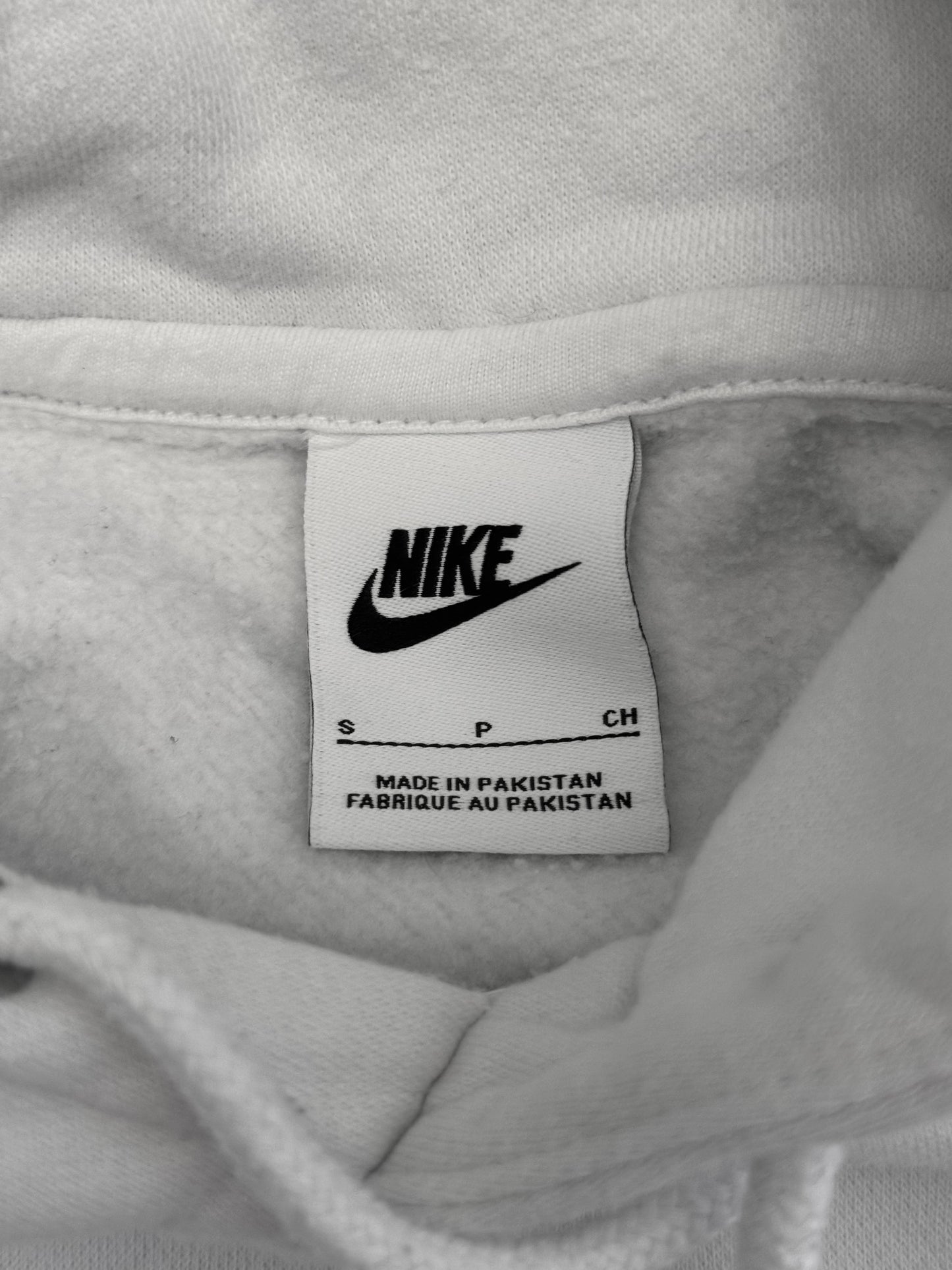 Close-up of a white Nike garment label sewn inside the collar of a light-colored hoodie, with the Nike swoosh and text reading 'MADE IN PAKISTAN' visible.