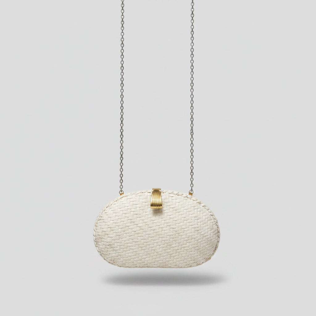 A small woven clutch bag in a cream or off-white color with a gold-tone clasp, hanging from a silver chain. The clutch is resting on a patterned beige and gray rug in an indoor setting.
