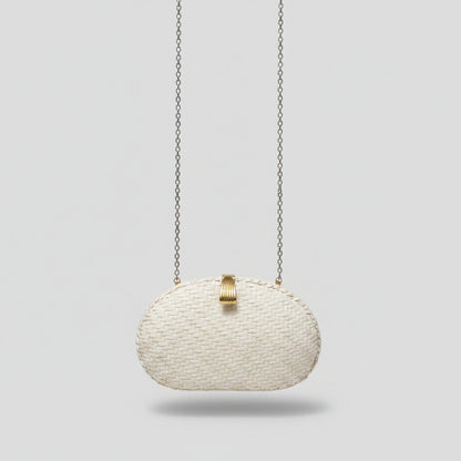 A small woven clutch bag in a cream or off-white color with a gold-tone clasp, hanging from a silver chain. The clutch is resting on a patterned beige and gray rug in an indoor setting.