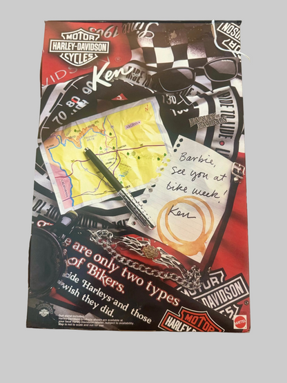 A Harley-Davidson themed notebook or journal cover featuring a collage of logos, a map, a pen, sunglasses, and handwritten notes, photographed against a light tiled floor.
