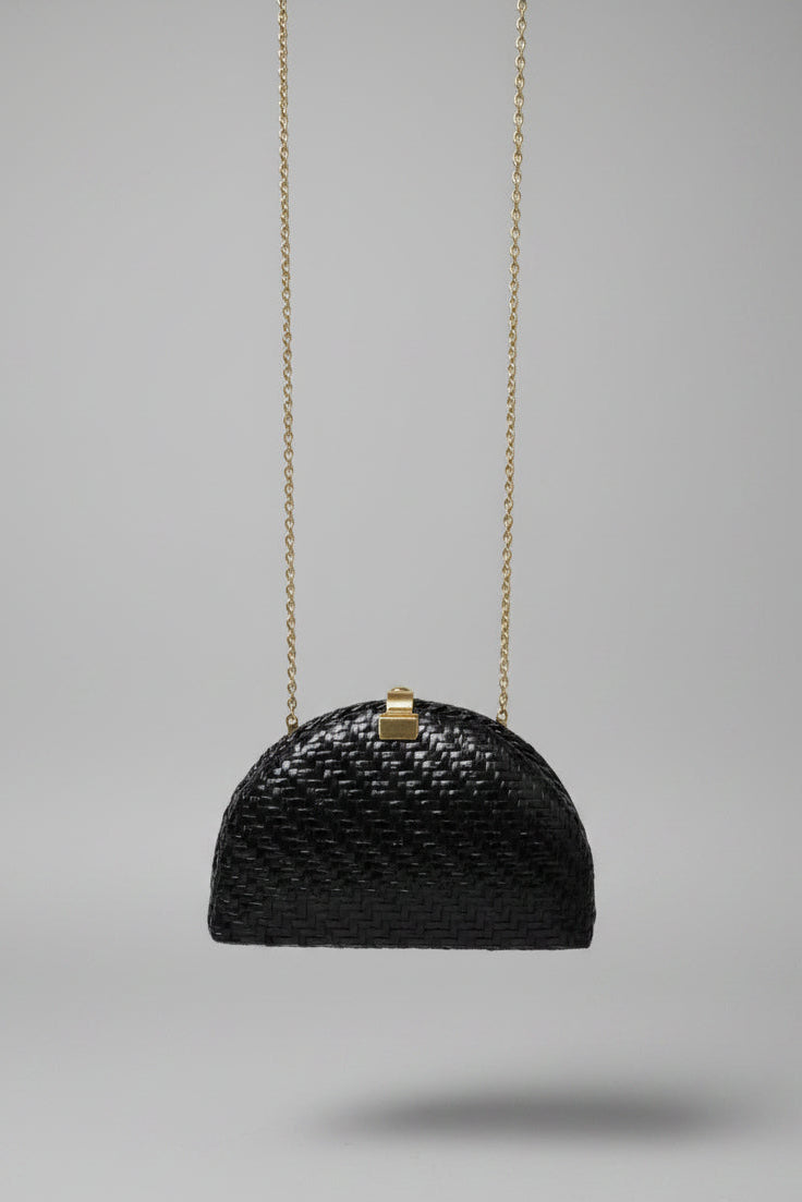 A small black woven handbag with a gold clasp and gold chain strap, photographed hanging in an indoor setting with a patterned rug and furniture visible in the background.