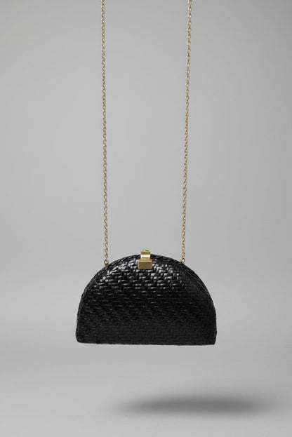 A small black woven handbag with a gold clasp and gold chain strap, photographed hanging in an indoor setting with a patterned rug and furniture visible in the background.
