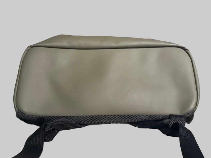 A light olive green waterproof pouch or small bag with black straps, resting on a textured white blanket in a home setting with framed pictures on the wall.