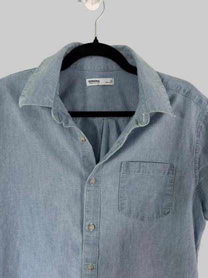 A light blue chambray button-down shirt by Domina, hanging on a black hanger against a white paneled door.
