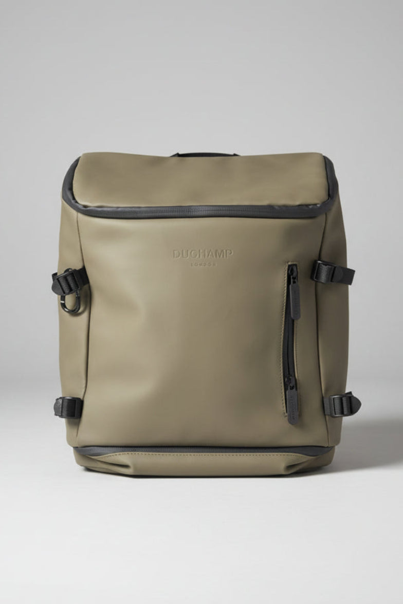A beige leather backpack with dark gray trim and zippers, featuring the brand name 'DUCHAMP' embossed on the front, resting on a textured blanket in a bedroom setting.