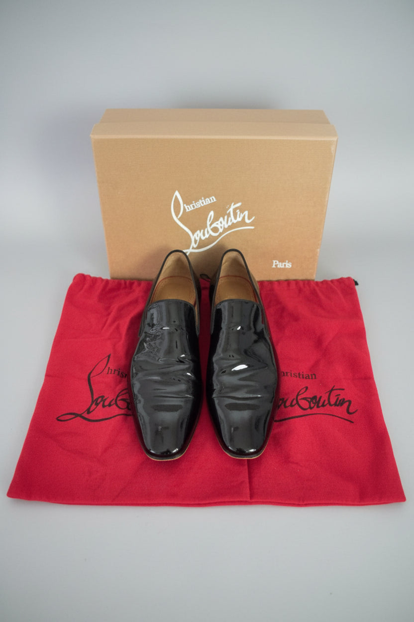 Black patent leather shoes on a red Christian Louboutin mat with a branded box in the background.