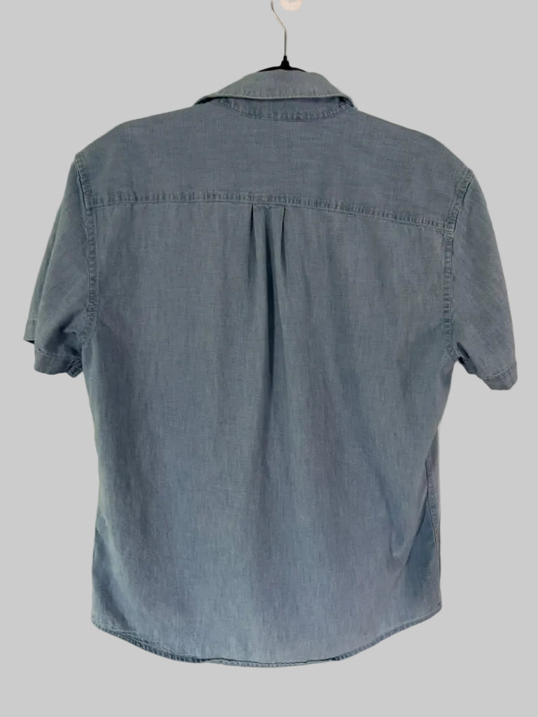 A light blue denim short-sleeve button-up shirt hanging on a black hanger against a white paneled door.