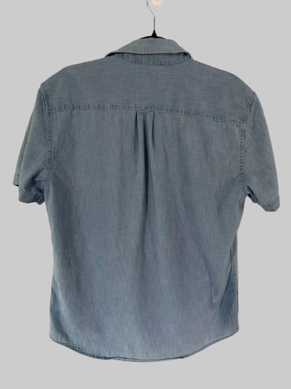 A light blue denim short-sleeve button-up shirt hanging on a black hanger against a white paneled door.