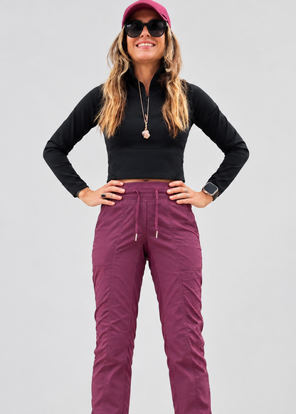 A woman modeling high-waisted, drawstring cargo pants in a dusty purple/magenta color, paired with a black long-sleeve top, sunglasses, and a pink cap, against a solid gray studio background.