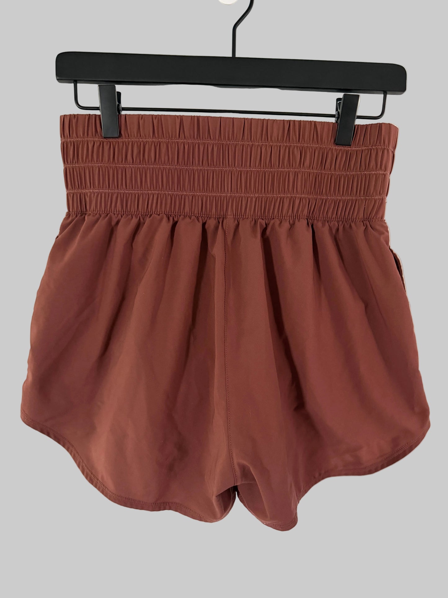 A pair of rust-colored, high-waisted shorts with an elasticized waistband, hanging from a black hanger against a white paneled door.