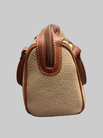 A small pebbled leather handbag in beige and brown, photographed standing on a dark wood floor in front of window blinds.