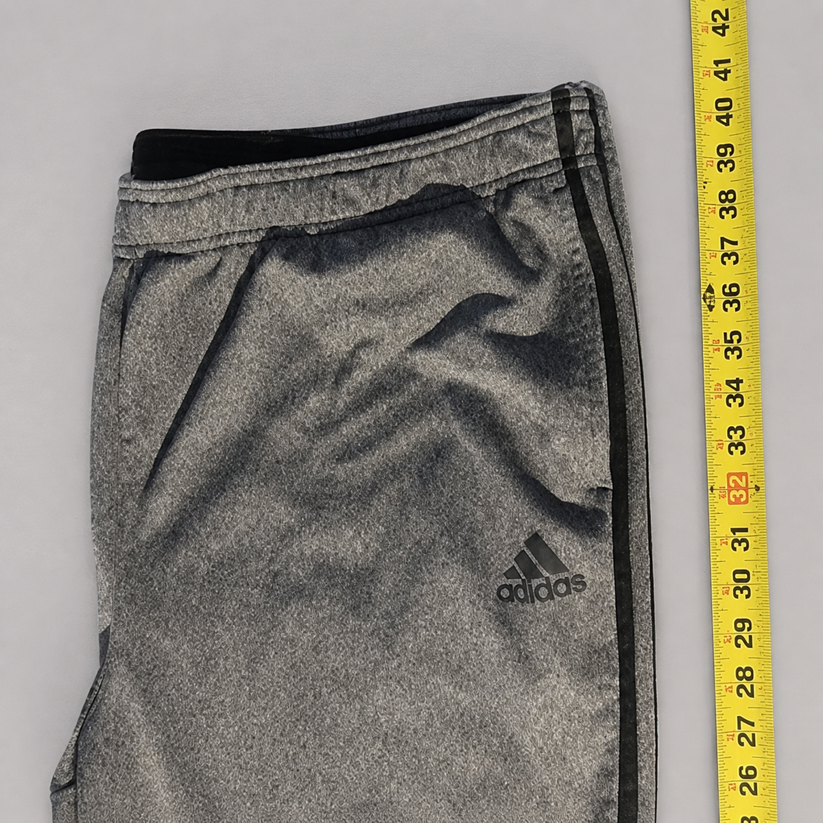 Gray heathered athletic shorts with a black side stripe and the Adidas logo, shown next to a yellow measuring tape on a light gray background.